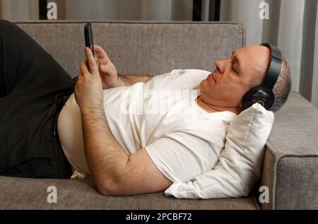 Un uomo di mezza età si trova sul divano, ascolta la musica con le cuffie, guarda il telefono. Foto Stock
