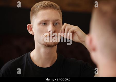 Ritratto di giovane uomo bello con baffi sul manubrio ricci e barba, ha un'acconciatura alla moda, indossa una t-shirt nera casual, guarda con fiducia nel mirr Foto Stock