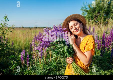 Ritratto di giovane donna che tiene bouquet di fiori lupino camminare nel prato estivo. Elegante ragazza raccolta fiori viola indossare cappello di paglia e godendo l Foto Stock