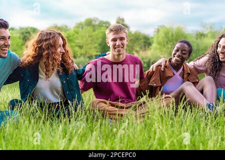 Un gruppo diversificato di amici abbraccia e sorride felicemente mentre si siede sull'erba in un parco - vita all'aperto, amicizia, integrazione razziale, fiducia e ca Foto Stock