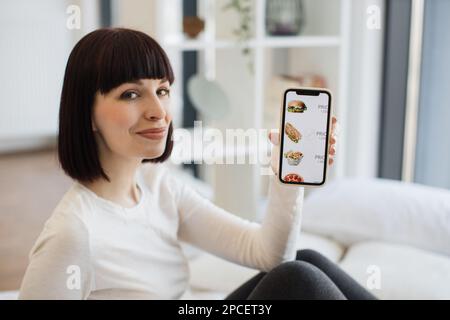 Donna caucasica piacevole in abbigliamento casual utilizzando moderno smartphone per ordinare fast food con sconto online mentre si soggiorna a casa. Concetto di persone, g Foto Stock