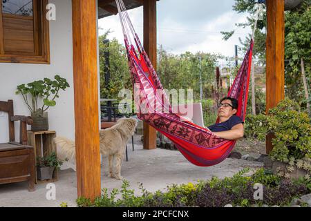 Latino uomo reclinato in un'amaca rossa che lavora sul suo laptop con un cane che lo accompagna Foto Stock