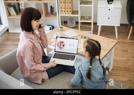 Bella donna caucasica che tiene un computer portatile e parla sullo smartphone mentre si siede con una bella figlia sul divano. Famiglia felice di due fare online ordine di pizza con sconto del 65% Foto Stock