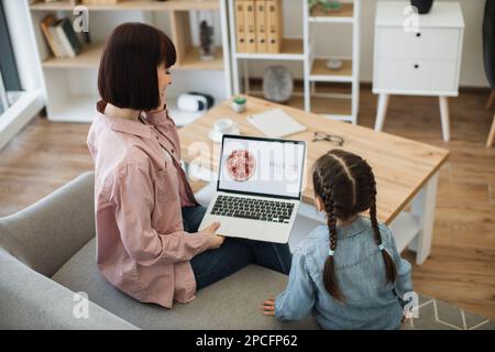 Bella donna caucasica che tiene un computer portatile e parla sullo smartphone mentre si siede con una bella figlia sul divano. Famiglia felice di due fare online ordine di pizza con sconto del 65% Foto Stock