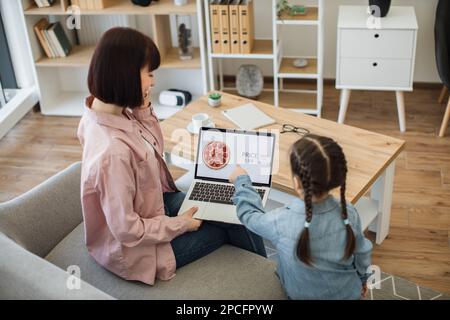 Bella donna caucasica che tiene un computer portatile e parla sullo smartphone mentre si siede con una bella figlia sul divano. Famiglia felice di due che fanno orde in linea Foto Stock