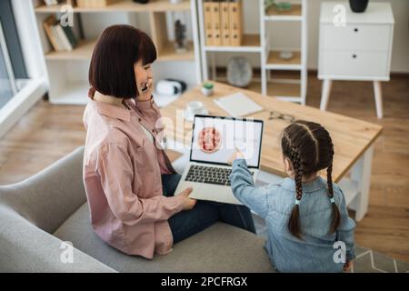 Bella donna caucasica che tiene un computer portatile e parla sullo smartphone mentre si siede con una bella figlia sul divano. Famiglia felice di due che fanno orde in linea Foto Stock