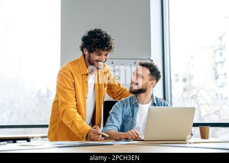 Due dipendenti maschi creativi di successo e amichevoli lavorano insieme a progetti in un ufficio moderno, discutendo nuove idee interessanti, progettando scadenze e strategia di promozione Foto Stock