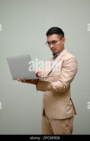 Professionista e stressato uomo d'affari asiatico millenario in tuta formale d'affari utilizzando il suo portatile, in piedi contro uno sfondo verde studio. Foto Stock