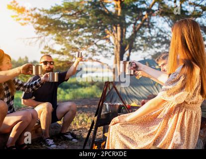 La compagnia di ragazze e ragazzi festeggia una vacanza nella natura. Stringono i bicchieri con le bevande. Riposatevi in estate nella foresta sulle rive della riva del fiume Foto Stock