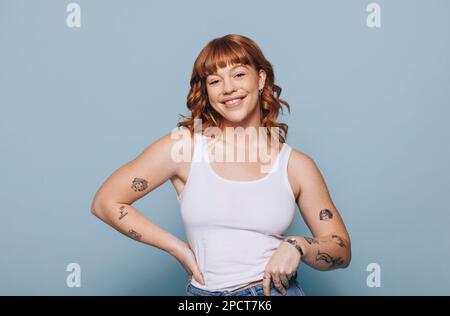 Giovane donna spensierata con tatuaggi sorridenti alla fotocamera mentre si è in piedi in uno studio. Ritratto di una giovane donna felice con i capelli di zenzero in piedi contro Foto Stock