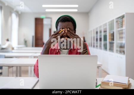 Esausto studente nero ragazzo sedersi in biblioteca con notebook sensazione malessere strofinare gli occhi prova concentrato Foto Stock
