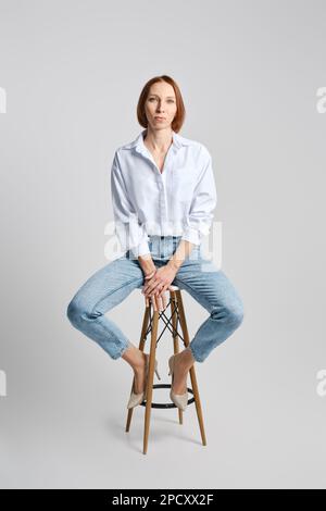 Ritratto di stile di vita di donna di mezza età caucasica a testa rossa in camicia bianca e jeans blu seduti su uno sgabello alto Foto Stock