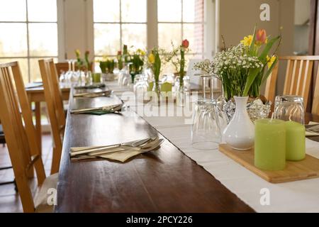 Grande tavolo in legno decorato per una cena con fiori primaverili, candele verdi e vari bicchieri da bere su un tavolo leggero corridore in una sala da pranzo ro Foto Stock