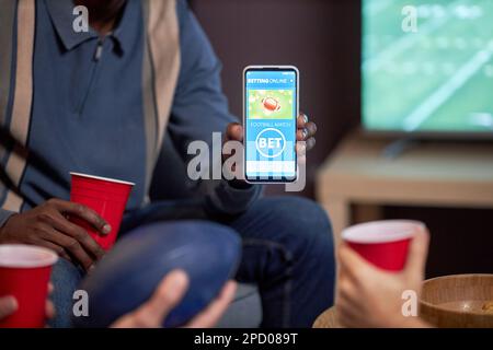 Primo piano di un uomo che tiene uno smartphone con l'app scommesse sportive alla fotocamera mentre guarda la partita di calcio a casa, copia spazio Foto Stock