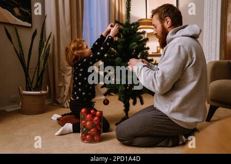 Zenzero padre e figlio in abbigliamento casual decorazione albero di Natale in accogliente soggiorno Foto Stock