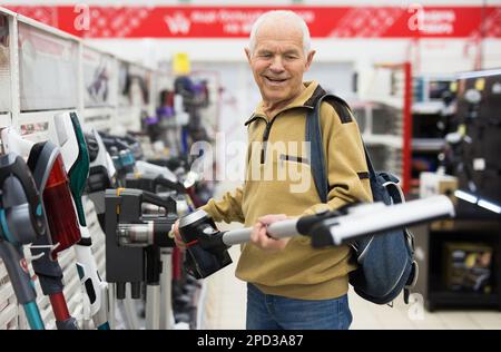 Uomo anziano in grigio pensionato guardando verticale aspirapolvere hoover al banco in showroom di elettrodomestici ipermercato reparto Foto Stock