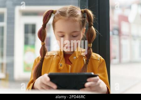 Ragazza bianca cute guardare video online con uno smartphone. Ritratto di adorabile bambino di 9 anni che gioca gioco mobile su un gadget Foto Stock