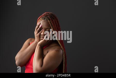 Una donna afroamericana con trecce rosse che le ricoprono il volto con le mani su uno sfondo scuro Foto Stock