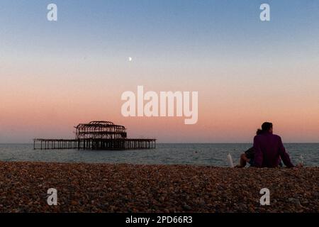 Giovani coppie guardano il tramonto sulla spiaggia di Brighton con il Molo Ovest e la luna sullo sfondo - Brighton & Hove, East Sussex, UK Foto Stock