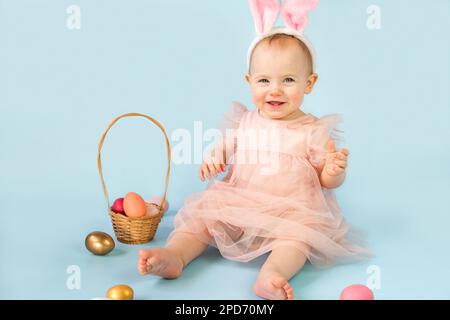 Ragazza felice, dieci mesi, con archetto per orecchie conigliate, seduta su sfondo azzurro dello studio e giocando con uova di pasqua colorate. Piccolo Chi Foto Stock