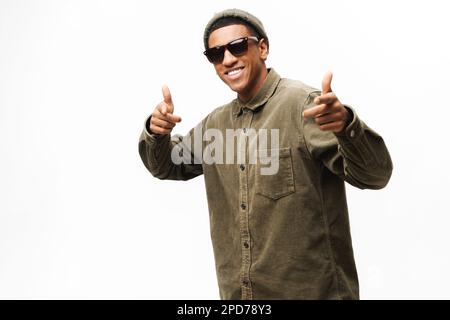 Giocoso afro-americano in occhiali da sole, cappello alla moda e camicia che indica la macchina fotografica isolato su bianco, positivo uomo allegro godendo di divertirsi, scegliendo voi, fingendo di fare un colpo da immaginate pistole Foto Stock