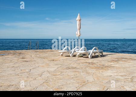 Autunno con sedie a sdraio vuote di plastica bianca accanto all'ombrellone a strisce blu e bianche sulla costa della Croazia, in Europa Foto Stock