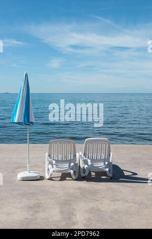 Autunno con sedie a sdraio vuote di plastica bianca accanto all'ombrellone a strisce blu e bianche sulla costa della Croazia, in Europa Foto Stock