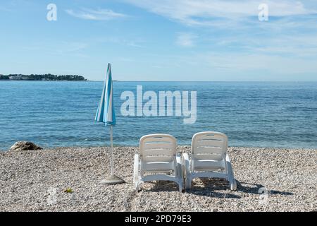 Autunno con sedie a sdraio vuote di plastica bianca accanto all'ombrellone a strisce blu e bianche sulla costa della Croazia, in Europa Foto Stock