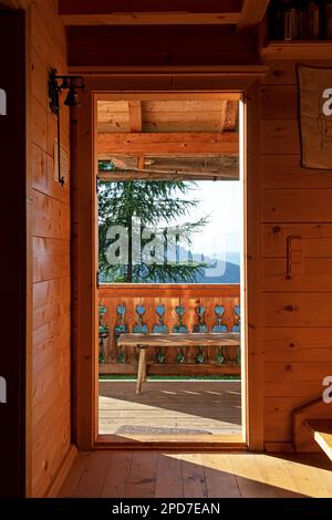 vista attraverso la porta aperta della capanna alpina in legno verso ringhiere in legno scolpite e paesaggio alpino alla luce del sole d'estate Foto Stock