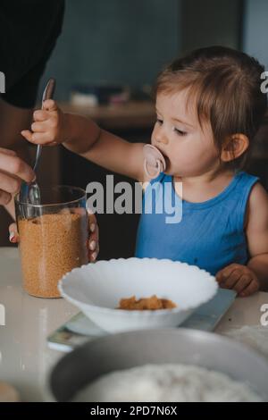 Vista ravvicinata della bambina che aggiunge farina con cucchiaio in un piatto preparando biscotti insieme alla madre. I bambini mescolano la farina nella ciotola, aggiungono Foto Stock