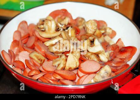 Funghi e salsicce vengono fritti in una padella rossa su una stufa a gas, ripieno per la pizza Foto Stock