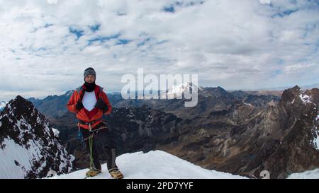 Giovane arrampicatore maschio in piedi sulla cima della montagna innevata indossando casco, faro, cappotto rosso e corda durante una giornata nuvolosa Foto Stock