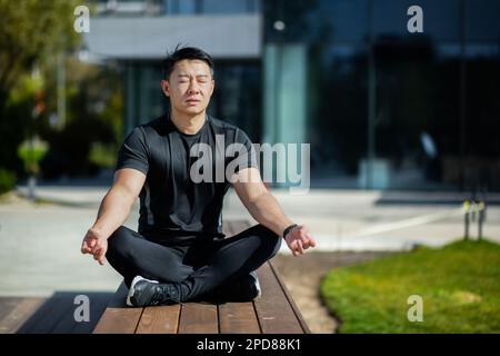 L'uomo asiatico medita nella posizione del loto, l'uomo nel parco vicino al lago esegue esercizi di yoga, lo sportivo si riposa con un mento chiuso dopo esercizi fisici attivi e corsa. Foto Stock