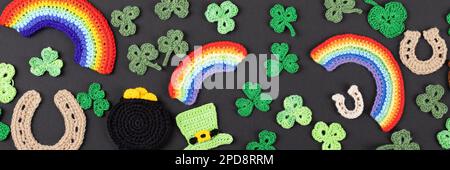 Concetto del giorno di San Patrizio. Composizione a maglia di un cappello verde, una pentola d'oro, un ferro di cavallo, un arcobaleno e shamrock verde su sfondo nero. Banner Foto Stock