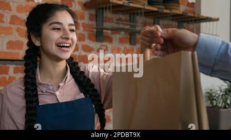 Amichevole giovane donna indiana cameriera barista che dà il sacchetto di cibo da asporto al cliente venditore femmina cafe dipendente in grembiule in attesa di consegna di ordine da asporto Foto Stock