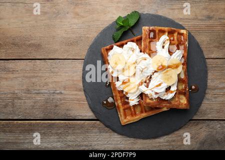 Piatto di ardesia di deliziosi waffle belgi con banana, panna montata e salsa al caramello su tavolo di legno, vista dall'alto. Spazio per il testo Foto Stock