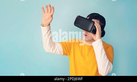Giovane uomo casual che usa gli occhiali VR, tocca qualcosa di invisibile nello spazio copia, uomo millenario che vive l'avventura in realtà virtuale sullo studio blu Foto Stock