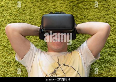 Il giovane uomo caucasico giace su un pavimento nel soggiorno con gli occhiali VR con il sorriso sul viso. visualizzazione di video 360 Foto Stock