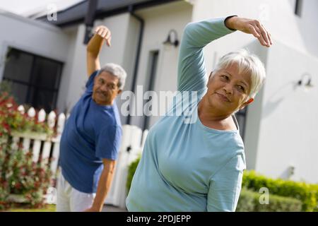 Coppia anziana biraciale con braccio sollevato facendo stretching mentre in piedi in cortile contro casa Foto Stock