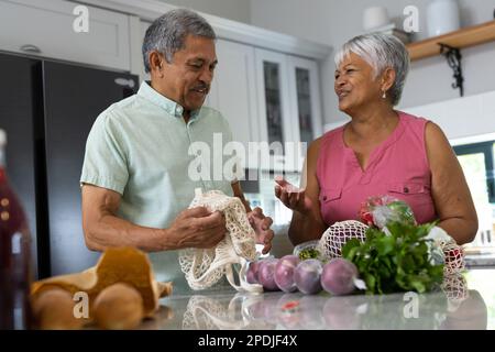 L'uomo anziano biraciale assiste la moglie nello sgombrare i generi alimentari dai sacchetti della maglia sul banco della cucina nel paese Foto Stock