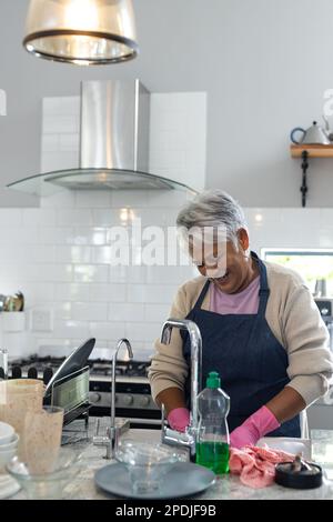 Donna anziana sorridente biraciale che indossa piatti di pulizia grembiule in cucina lavello, spazio copia Foto Stock