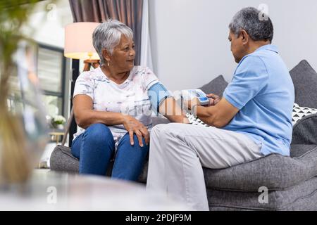 Uomo anziano biraciale che controlla la pressione sanguigna della moglie con manometro mentre si siede sul divano a casa Foto Stock