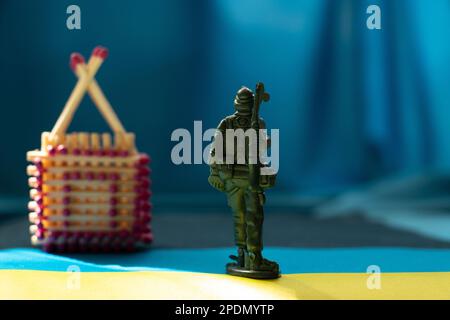 Una piccola casa di legno fatta di fiammiferi si trova sulle bandiere dell'Ucraina sotto il sole e un soldato di plastica dei bambini si trova di fronte alla casa, retur Foto Stock