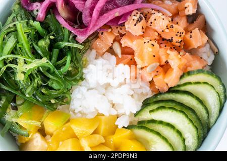 Ciotola di poke con riso, salmone, cetriolo, mango, cipolla, insalata di wakame, semi di papavero e semi di girasoli isolati su sfondo bianco Foto Stock
