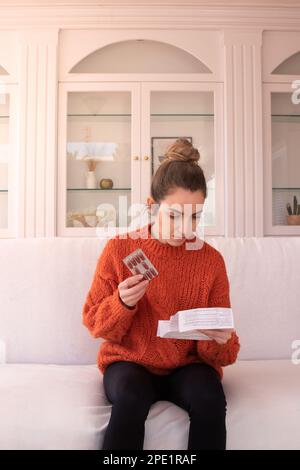 Giovane donna in maglia arancione maglione seduto sul divano bianco e leggere le istruzioni mediche a casa Foto Stock