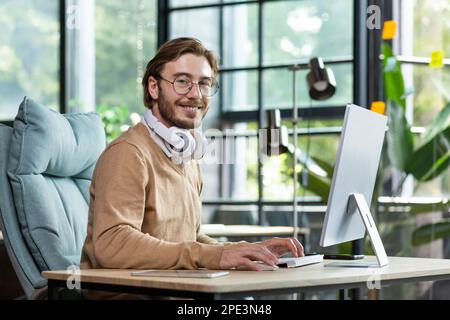 Ritratto di un giovane freelancer, programmatore siede in ufficio in cuffie bianche e lavora al computer. Sorridendo guardando la fotocamera. Foto Stock