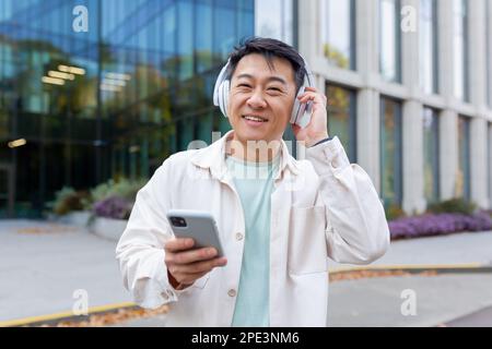 Giovane studente asiatico maschio in piedi fuori dal campus indossando cuffie. Tiene un telefono nelle mani. Ascolta musica, parla. Sorridendo guardando la fotocamera. Foto Stock