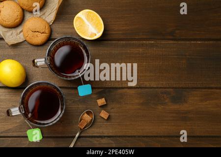 Bustine di tè in tazze di vetro di acqua calda, biscotti e cubetti di zucchero su tavolo di legno, piatto. Spazio per il testo Foto Stock