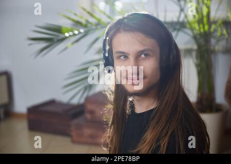 Ritratto di un giovane uomo con capelli lunghi che indossa le cuffie a casa. Foto Stock