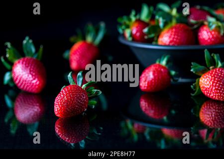 Una ciotola piena di fragole succose con alcune sparse su un tavolo di vetro di fronte. Foto Stock